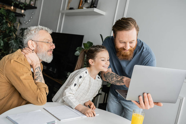 Cheerful kid doing homework near tattooed gay parents with laptop at home  - Photo, Image