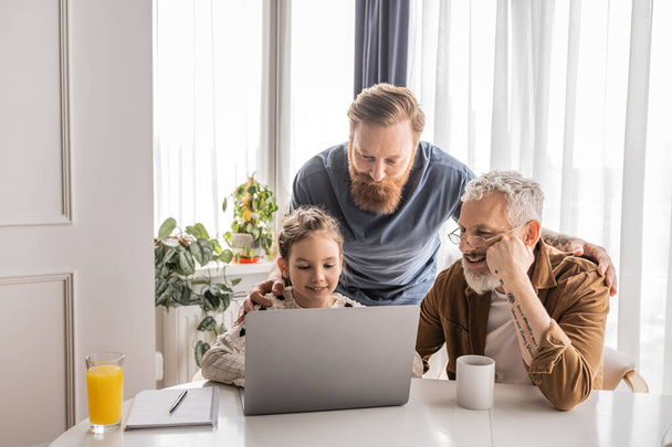 Smiling gay parents with daughter using laptop near notebook and drinks at home  - Valokuva, kuva
