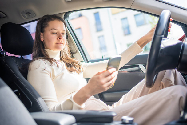Młoda kobieta siedząca w samochodzie i używająca smartfona, bizneswoman zajęta prowadzeniem. Wysokiej jakości zdjęcie - Zdjęcie, obraz