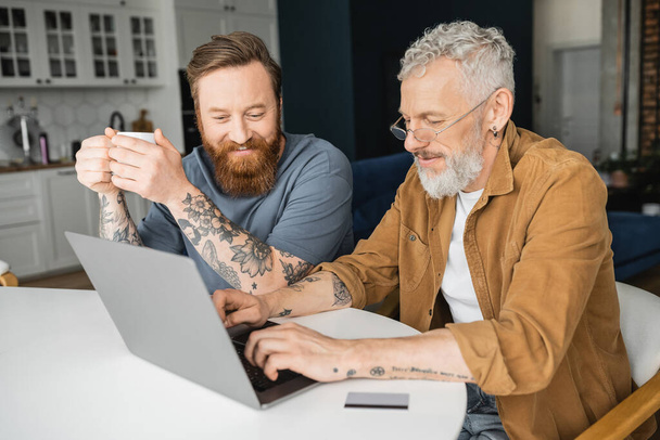 glimlachen gay man holding kopje koffie terwijl getatoeëerde partner met behulp van laptop in de buurt van credit card thuis  - Foto, afbeelding