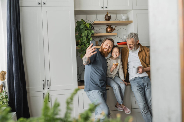 Hymyilevä homoseksuaalinen mies ottaa selfien tyttären ja kumppanin kanssa keittiössä  - Valokuva, kuva