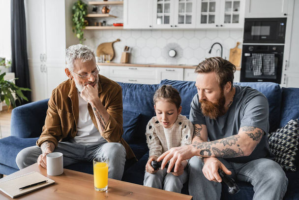KYIV, UKRAINE - APRIL 19, 2023: Tattooed gay dad helping daughter with joystick near partner and drinks at home  - Zdjęcie, obraz