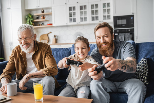 KYIV, UKRAINE - APRIL 19, 2023: Smiling gay parent playing video game with daughter near partner and drinks at home  - Foto, imagen