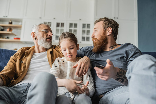 Gay parents with remote controller talking near preteen daughter at home  - Fotoğraf, Görsel