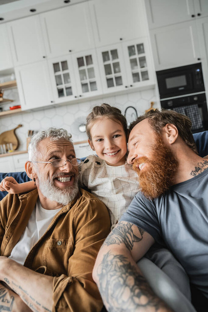 Positive tattooed same sex parents sitting near preteen daughter on couch at home  - Photo, Image