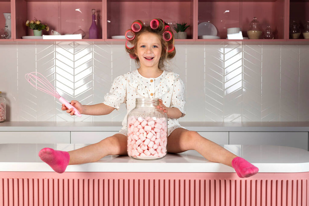 Ragazzina felice che indossa rulli per capelli e abito a pois bianco, si siede su un tavolo da cucina rosa, con un grande vaso di vetro con dolci marshmallow o caramelle.. - Foto, immagini