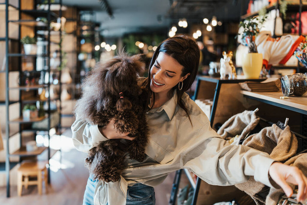 Beautiful young woman enjoying in modern pet shop together with her adorable brown toy poodle.  - Fotó, kép
