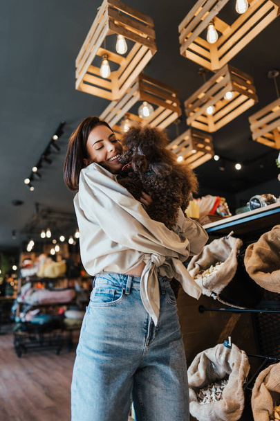 Beautiful young woman enjoying in modern pet shop together with her adorable brown toy poodle.  - Photo, image