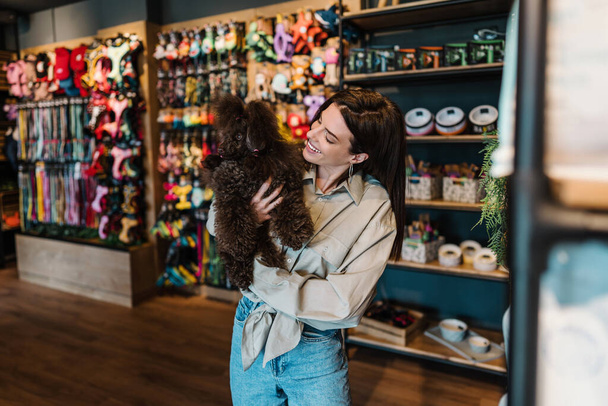 Beautiful young woman enjoying in modern pet shop together with her adorable brown toy poodle.  - Foto, Imagen