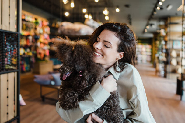 Beautiful and happy young woman sitting in modern pet shop cafe bar and enjoying in fresh coffee together with her adorable brown toy poodle.  - Foto, afbeelding