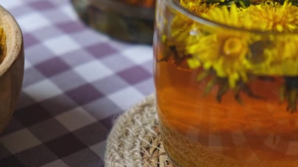 Gyermekláncfű virág egészséges tea üveg teáskanna és üveg csésze az asztalon. Ízletes gyógynövény tea ömlött friss pitypang virágok otthon nyári napon, közel a kerthez. Zöld tisztás Forró pitypang tea a - Felvétel, videó