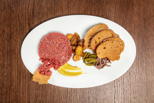 Veal tartare with rye bread toasts, fried potatoes, pickled cucumbers and anchovies with mustard sauce. Food lies on a light plate on a brown wooden tabletop. - Photo, Image