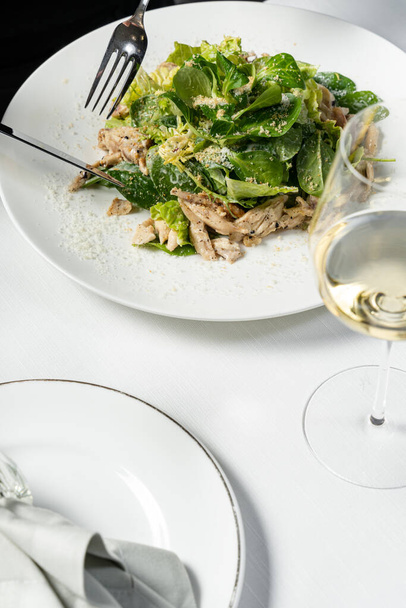 Mix salad with chicken breast and Parmesan cheese on a light ceramic plate. The plate is on a light tablecloth, next to it is a glass of white wine and cutlery. - Photo, image