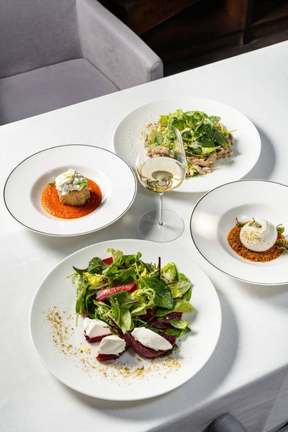 Mozzarella mit Gemüseeintopf und Auberginen paniert mit Stracciatella, Hühnersalat und glasiertem Rote-Bete-Salat. Gerichte liegen in hellen Keramiktellern auf einer hellen Tischdecke. - Foto, Bild