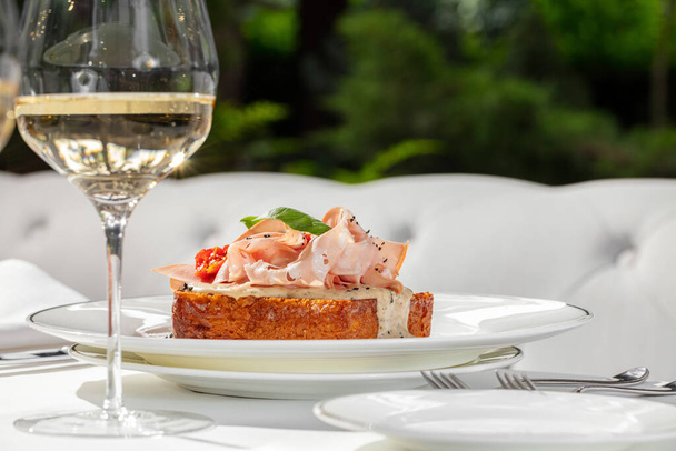 Brioche toast with ham and tomato walnut sauce, topped with basil leaves and herbs. The toast is in a light ceramic plate, a glass of white wine is next to it. - Photo, Image