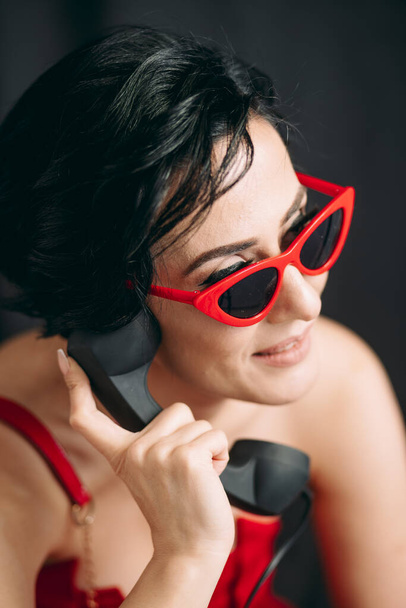 Divatos fiatal hölgy piros szemüvegben beszél telefonon előtt fekete háttér - Fotó, kép