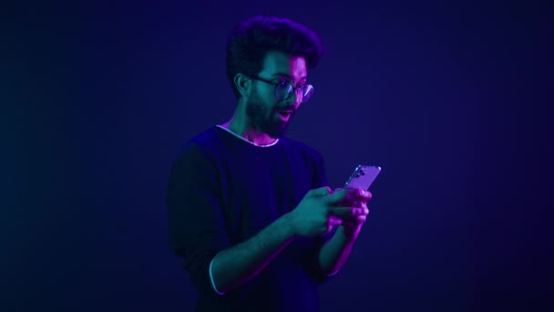 Indian man Arabian male model guy win mobile phone success victory smartphone achievement winning game cellphone internet betting online money prize at ultraviolet neon dark blue studio background - Footage, Video