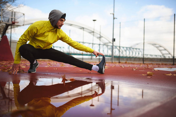 Joven atleta hombre estirando sus músculos antes de correr en el puente en un día lluvioso - Foto, imagen