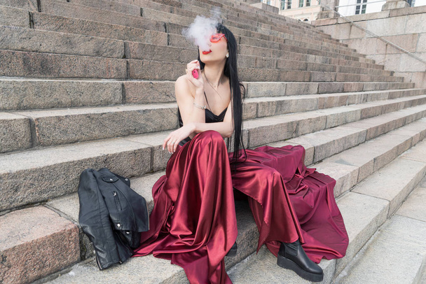 Uma mulher sexy e uma saia de cetim vermelho brilhante está sentado nos degraus. jaqueta de couro nas proximidades. Ela fuma um cigarro eletrônico. - Foto, Imagem