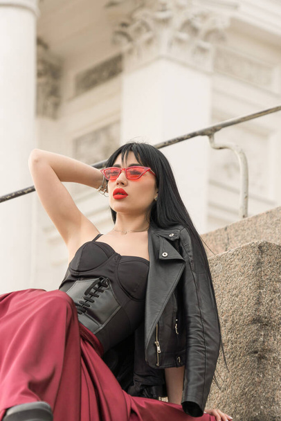 Woman with long hair, perfect figure, sexy. she is in a satin, long red skirt, leather jackets on the steps. Posing in the city. - Φωτογραφία, εικόνα