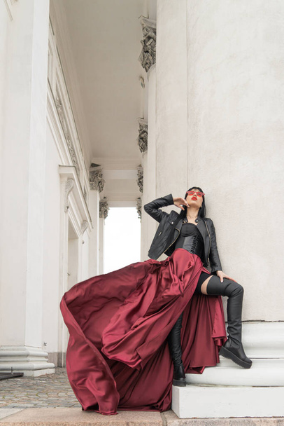 Woman with long hair, perfect figure, sexy. she is in a satin, long red skirt, leather jackets. Posing in the city at the tall ancient columns - Φωτογραφία, εικόνα