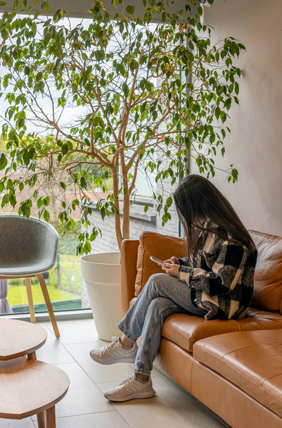One young caucasian brunette girl with flowing hair and with a mobile phone in her hands sits on a brown sofa in the waiting room in the dentistry, close-up side view. - 写真・画像