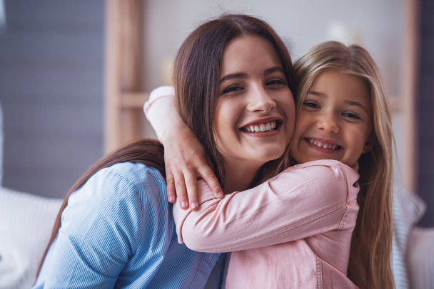 Portrait of beautiful young mother and her daughter hugging, looking at camera and smiling - Foto, imagen