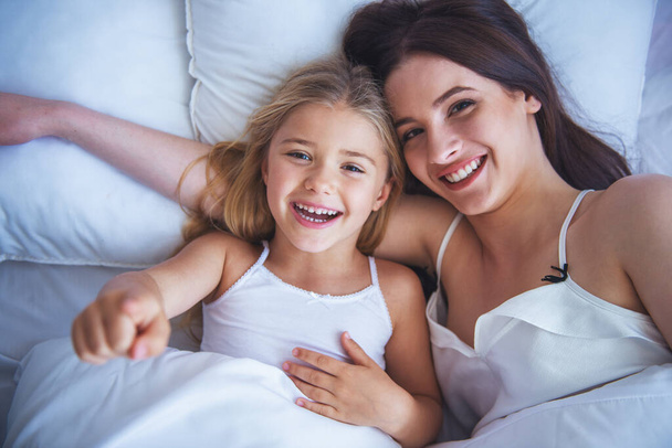 Top view of beautiful young mother and her daughter hugging, looking at camera and smiling while lying in bed at home - Photo, image