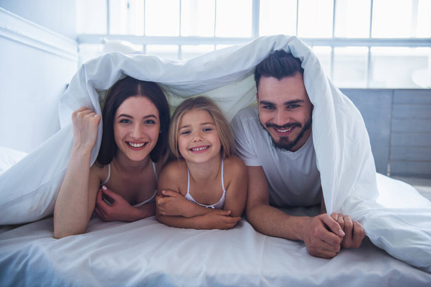 Retrato de una hermosa madre joven, padre y su hija mirando a la cámara y sonriendo mientras están acostados en la cama cubiertos con manta
 - Foto, Imagen