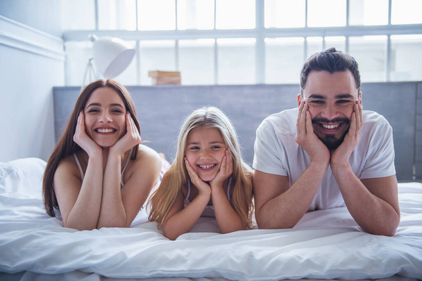 Retrato de una hermosa madre joven, padre y su hija mirando a la cámara y sonriendo mientras están acostados en la cama y apoyados en sus manos
 - Foto, Imagen