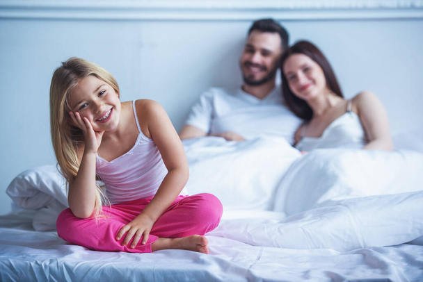 Nettes kleines Mädchen blickt in die Kamera und lächelt, während es auf dem Bett sitzt, ihre glücklichen Eltern im Hintergrund - Foto, Bild
