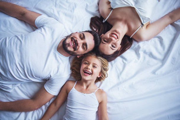 Top view of beautiful young mother, father and their daughter looking at camera and smiling while lying on bed head to head - Photo, Image