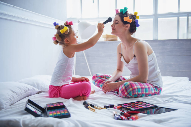 Mooie jonge moeder en haar dochter in pyjama en met haar krulspelden op bed thuis. Schattig klein meisje doet haar moeder make-up - Foto, afbeelding