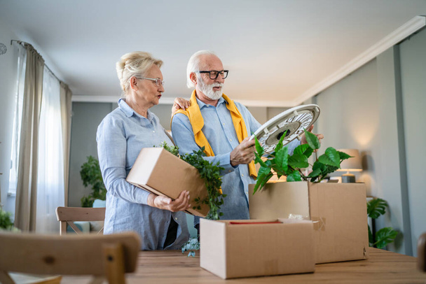 family senior couple man woman husband and wife pensioner grandfather grandmother moving in new apartment taking their stuff belongings and plants in or out of boxes packing or unpacking real people - Photo, Image