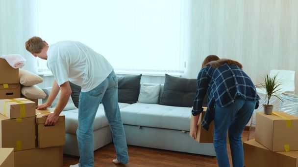 Rear of young couple man and woman entering the room carrying cardboard boxes on moving day and sit on sofa. Happy boyfriend and girlfriend moved in together in apartment. Slow motion. Home concept - 写真・画像