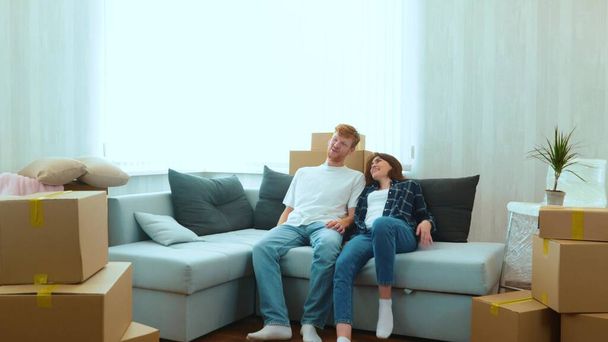 Cheerful Caucasian couple boyfriend and girlfriend moving in together in new house in good mood. Tired happy young married couple sitting down on sofa in new home with many boxes on moving day - 写真・画像