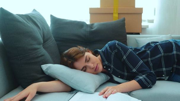 Beautiful young Caucasian woman lying on sofa in living room fallen asleep with book in new apartment with many cardboard boxes. Caring loving husband covers his wife with a blanket. Love concept - Zdjęcie, obraz