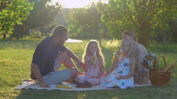 Caucasian family with cute blonde little girls in park in summer. Happy children are eating fruits sitting on blanket outdoor. Happy childhood. Family weekend. Kids eat bananas with parents on nature - Photo, image