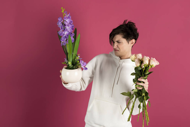Knappe jongeman in witte capuchon, met een keuze tussen een boeket rozen of bloemen en een plant in een pot. Een man die beslist over een cadeau voor een date. Geïsoleerd op roze. - Foto, afbeelding