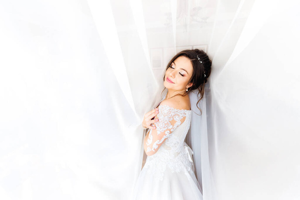 白いドレスの美しい花嫁は白いカーテンの背景に彼女の目を閉じました - 写真・画像