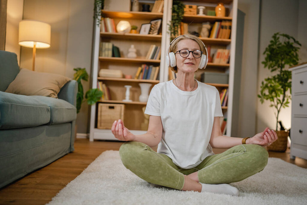 yksi nainen kypsä vanhempi valkoihoinen nainen käyttää kuulokkeita verkossa opastettu meditaatio harjoitellaan mindfulness jooga ilmentymä silmät kiinni kotona oikeita ihmisiä itsehoito käsite kopioi tilaa - Valokuva, kuva