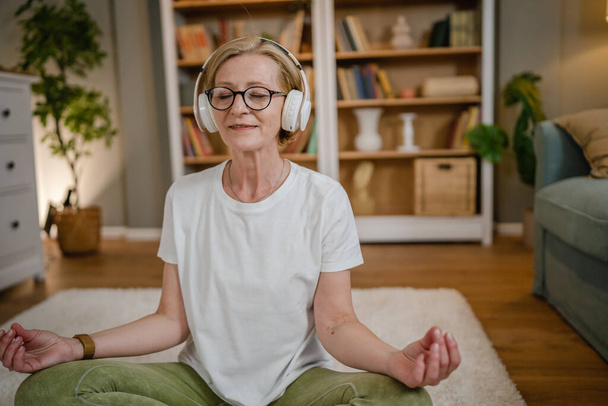 Yetişkin bir yaşlı beyaz kadın internet rehberli meditasyon için kulaklık kullanıyor. Farkındalık yogası yapıyor. Evde gözleri kapalı. - Fotoğraf, Görsel