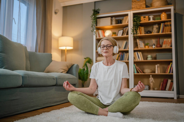 una mujer madura mayor caucásica femenina usando auriculares para la meditación guiada en línea practicando mindfulness manifestación de yoga con los ojos cerrados en casa personas reales autocuidado concepto copia espacio - Foto, Imagen