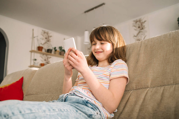 Uma adolescente senta-se no sofá em casa e usa um telefone celular, conversando e chamando amigos. Adolescentes e tecnologia.  - Foto, Imagem