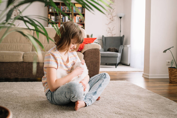 Una adolescente se sienta en el suelo en la sala de estar y juega con su amado gatito. El niño abraza y acaricia a la mascota. Niños y gatos - Foto, Imagen