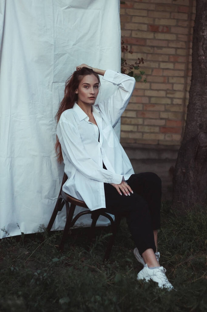 Modern photo of a girl in a white shirt against a white sheet. Posing on a chair. Laconic photos of a confident woman. Plant background - Foto, Imagem