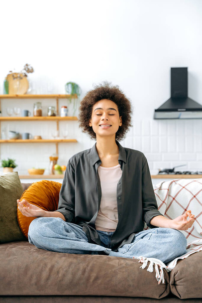 Függőleges fotó nyugodt, nyugodt, nyugodt afro-amerikai nőről alkalmi ruhában, otthon ül egyedül a nappaliban a kanapén, pihen és meditál lótuszpozícióban, mosolyog - Fotó, kép