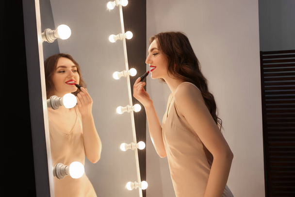 Beautiful young woman in elegant dress applying lipstick near mirror indoors - Foto, afbeelding