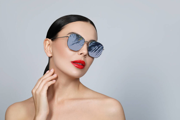 Attractive woman in stylish sunglasses on light grey background. Palm leaves and sky reflecting in lenses. Space for text - Φωτογραφία, εικόνα