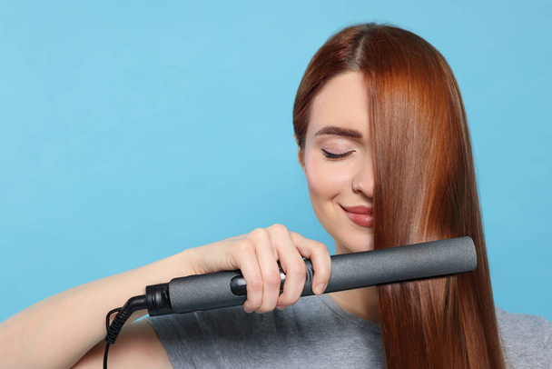 Beautiful woman using hair iron on light blue background, space for text - Foto, Imagen